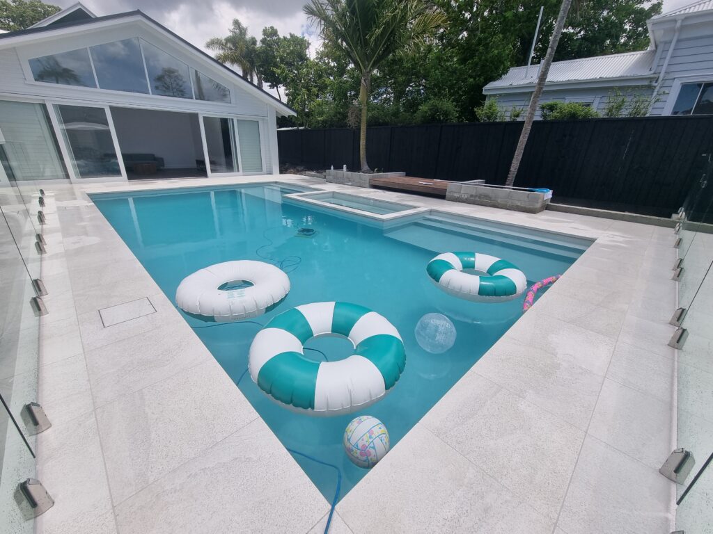 Pale stone pool coping around an Auckland pool with a glass pool fence